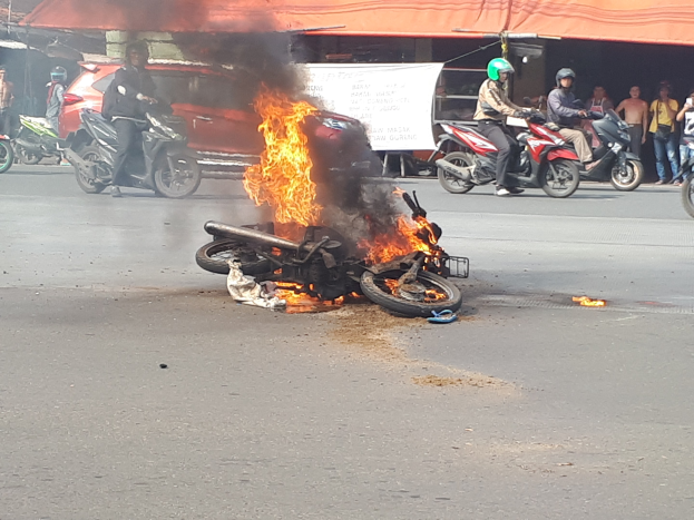 Motorrad in Flammen auf der Straße mit einem Auto und einem Van im Hintergrund, umgeben von Menschen in Helmen, einer Fahne mit Text und einem Zelt.