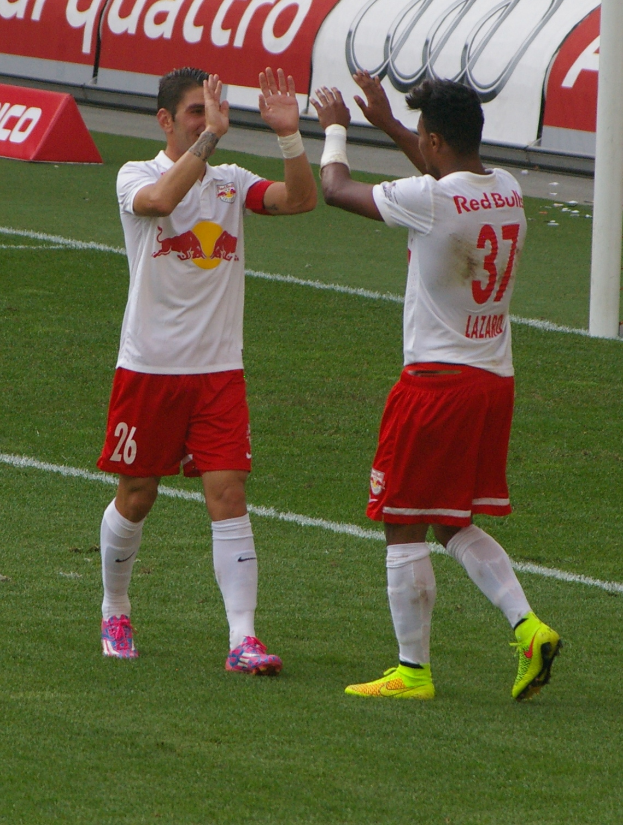 Zwei Fußballspieler klatschen sich auf einem Rasenfeld ab, im Hintergrund ein Pfosten und eine Tafel mit Text.