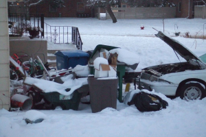 Eine Außenansicht mit Müll in der Mitte, einem Auto rechts, Treppen links, Bäumen oben und Eis am Boden.