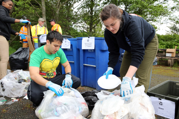Eine Gruppe von Menschen mit Handschuhen sammelt Müll in einem Park, umgeben von Plastikabdeckungen, Flaschen und anderem Schutt, mit einem Mülleimer und einer Holzbank rechts und Bäumen und einem klaren Himmel im Hintergrund.