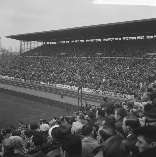 Schwarzes und weißes Foto eines vollen Fußballstadions mit Zuschauern, die ein Spiel verfolgen, mit Bannern, Pfählen, einer Hütte, Bäumen, einem Turm und einem bewölkten Himmel.