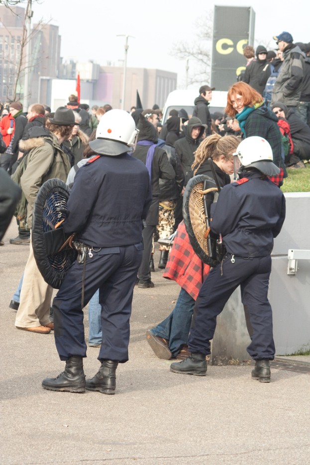 Eine Gruppe von Menschen, die auf einer Straße gehen, mit zwei Personen in der Mitte, die wie Polizisten aussehen, Gebäude im Hintergrund und Boden unten.