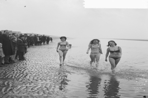 Eine Gruppe von Frauen in Bikinis, die an einem Strand entlanggehen, mit ein paar Menschen auf der linken Seite, dargestellt in Schwarz-Weiß.
