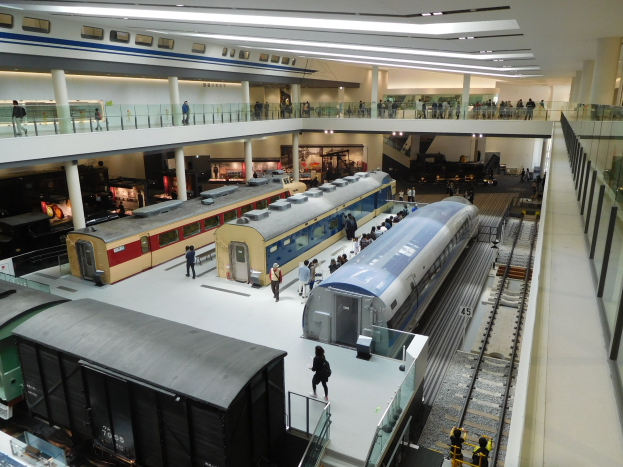 Ein großer Museumsraum mit verschiedenen Zügen in unterschiedlichen Größen und Farben, die von Menschen auf dem Boden, Geländern und Säulen betrachtet werden, unter Deckenbeleuchtung.