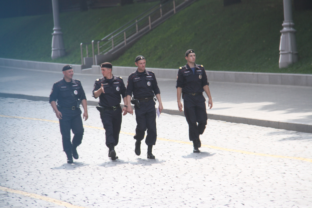 Drei Polizisten in Uniform und Mützen gehen eine Straße entlang mit Polen, Geländern, Treppen und Gras im Hintergrund.