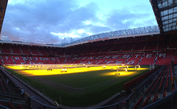 Old Trafford Stadium, Heimat von Manchester United, mit einem grünen Feld, umgeben von Sitzplätzen, hellen Lichtern, einer hüttenähnlichen Struktur auf der rechten Seite und einem Himmel mit weißen Wolken.