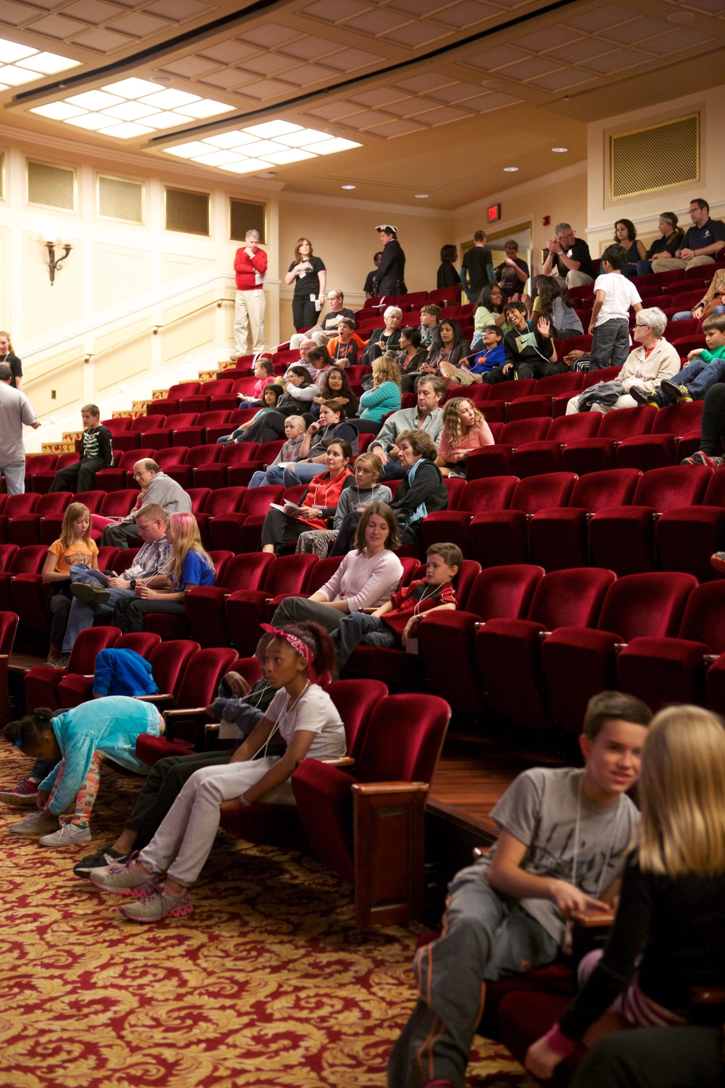 Eine große Gruppe von Menschen, darunter Kinder, die auf Stühlen sitzen und stehen, mit verstreuten Stühlen, Wand- und Deckenlampen im Hintergrund.