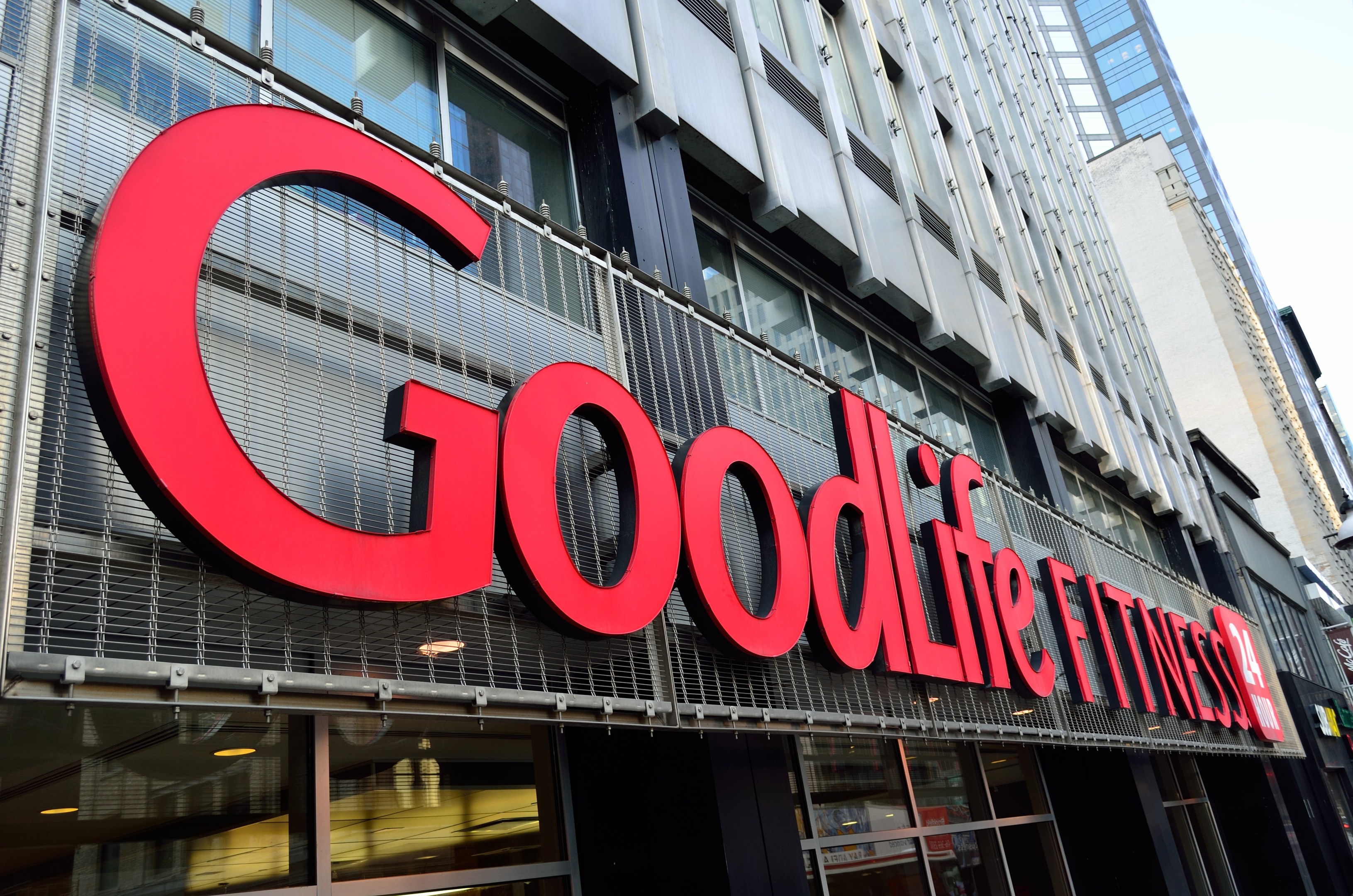 Außenansicht eines Goodlife Fitness Studios in New York City mit Glaswänden und einem Namensschild, umgeben von anderen Gebäuden und dem Himmel im Hintergrund.