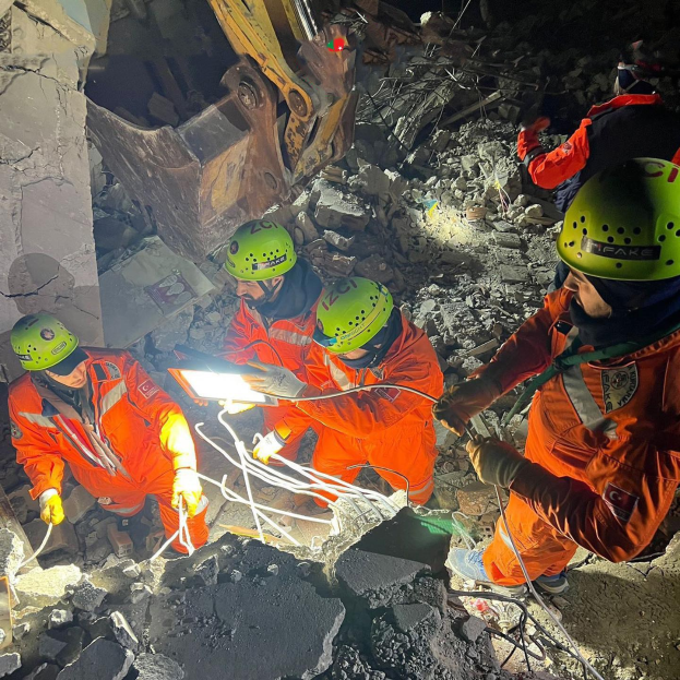 Gruppe von Menschen in orangen Uniformen und grünen Helmen bei der Arbeit an Schutt von einem eingestürzten Gebäude, mit Werkzeugen und Materialien, mit einem Kran im Hintergrund.