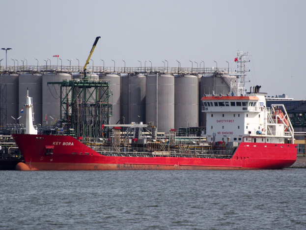 Ein großer roter und weißer Öltanker schwimmt auf einem Gewässer mit einer Brücke, Metallcontainern, Pfählen und einem klaren blauen Himmel im Hintergrund.
