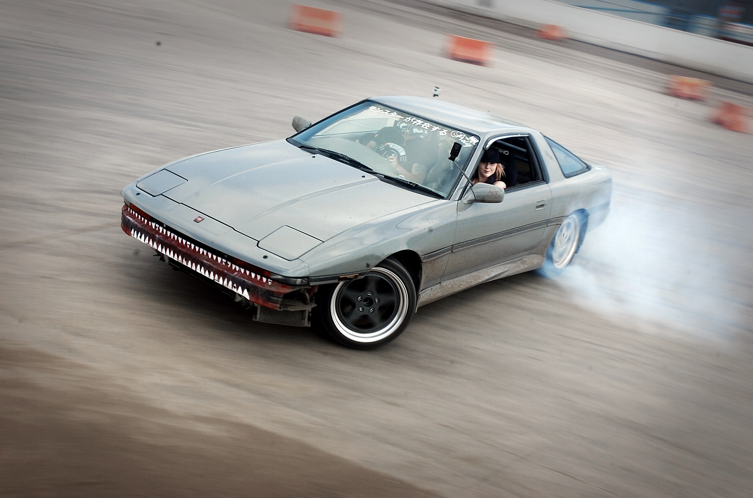 Eine Person mit Helm fährt ein Auto auf einer Strecke, aus dem Rauch austritt, umgeben von orangefarbenen Gegenständen, mit einem unscharfen Hintergrund, der hohe Geschwindigkeit andeutet.