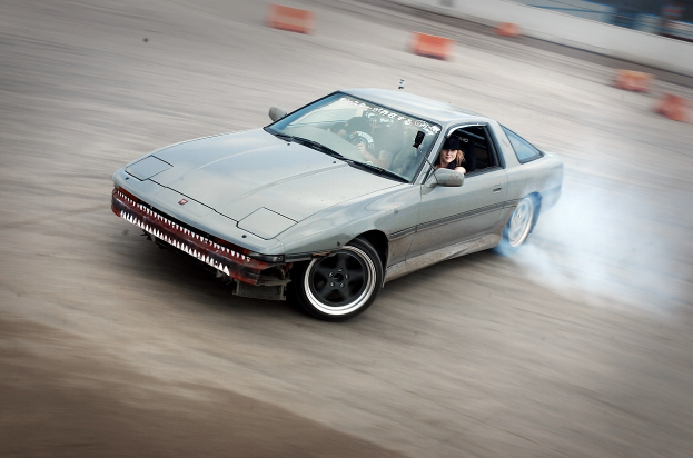 Eine Person mit Helm fährt ein Auto auf einer Strecke, aus dem Rauch austritt, umgeben von orangefarbenen Gegenständen, mit einem unscharfen Hintergrund, der hohe Geschwindigkeit andeutet.