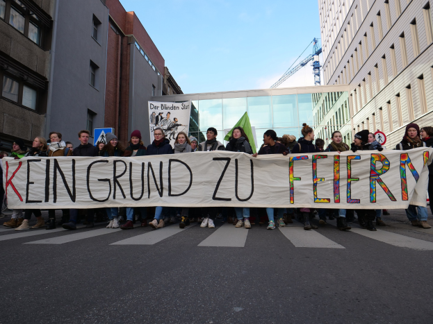 Demonstranten halten ein Transparent mit der Aufschrift "Kein Grund zu Feiern" gegen Sparmaßnahmen, mit Gebäuden, einem Schild, einem weiteren Transparent, einem Kran und einem bewölkten Himmel im Hintergrund.