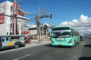 Ein grüner Bus fährt eine Straße mit hohen Gebäuden entlang, mit Fußgängern mit Regenschirmen und Strommasten mit Drähten im Vordergrund, unter einem bewölkten Himmel.