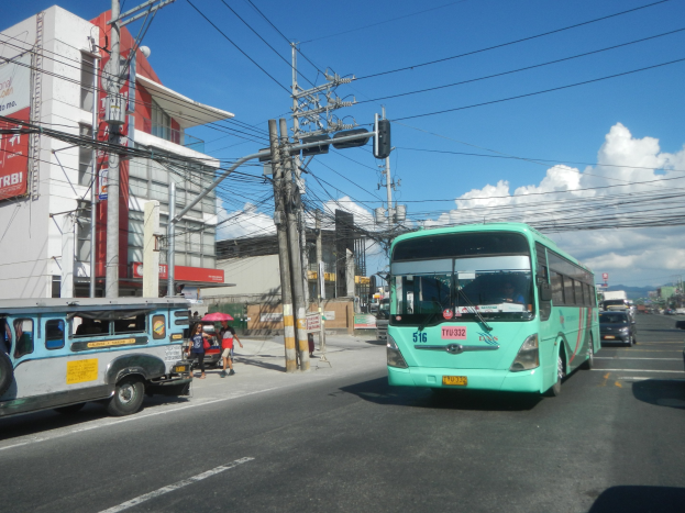 Ein grüner Bus fährt eine Straße mit hohen Gebäuden entlang, mit Fußgängern mit Regenschirmen und Strommasten mit Drähten im Vordergrund, unter einem bewölkten Himmel.