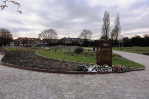 Eine Gedenkstätte im Park mit einem Backsteinweg, umgeben von Grün, Bänken und Bäumen, mit Gebäuden und einem bewölktem Himmel im Hintergrund und einer Tafel mit Text auf der rechten Seite.