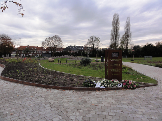 Eine Gedenkstätte im Park mit einem Backsteinweg, umgeben von Grün, Bänken und Bäumen, mit Gebäuden und einem bewölktem Himmel im Hintergrund und einer Tafel mit Text auf der rechten Seite.