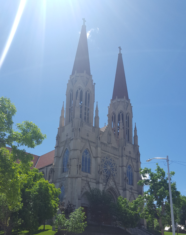Eine große Kirche mit zwei Türmen in einem Park, umgeben von Bäumen, Straßenmasten, Straßenlaternen, Strommasten, Stromkabeln, Treppen, Geländern und Gras unter einem klaren blauen Himmel.