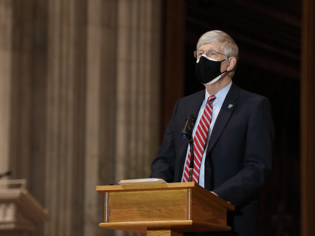Ein Mann im Anzug und mit Krawatte trägt eine Gesichtsmaske und steht an einem Podium mit einem Mikrofon und einem Buch, während er eine Rede hält. Im Hintergrund sind Säulen und eine Wand zu sehen, sowie ein weiteres Podium links von ihm.