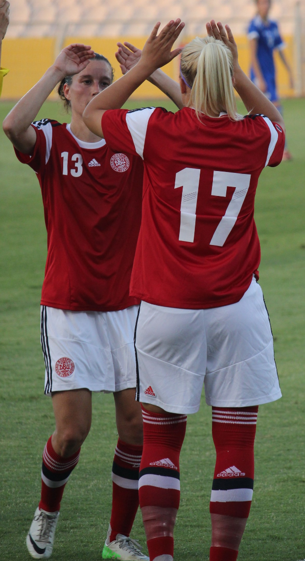 Zwei Frauen in roten T-Shirts und weißen Shorts klatschen auf einem Fußballfeld, mit einer Person in einem blauen Kleid und einer gelben Tafel im Hintergrund.