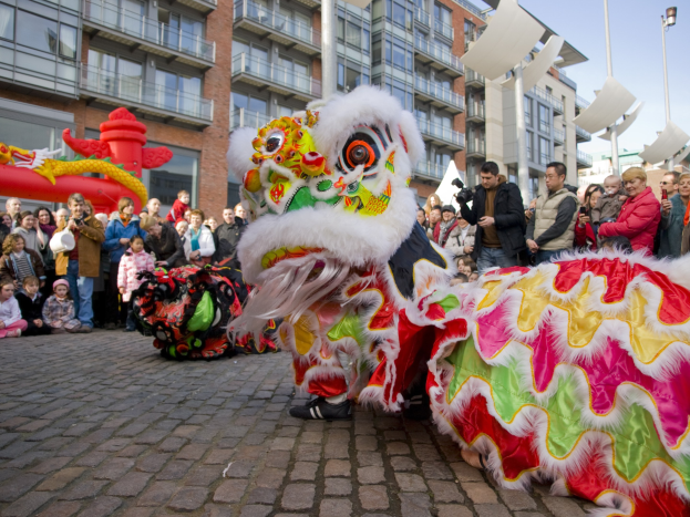 Ein farbenfrohes chinesisches Neujahrsfest in Amsterdam mit einem Löwen tanzen und einer Menge Schaulustiger, einige mit Kameras, vor einem Hintergrund aus Gebäuden, Laternenmasten und einem klaren blauen Himmel.