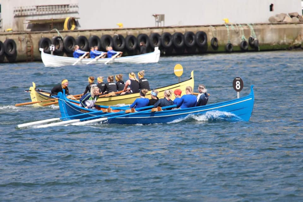 Eine Gruppe von Menschen in einem blauen und gelben Boot auf dem Wasser, die Paddel halten, mit einer Reifenwand und einem Gebäude im Hintergrund.