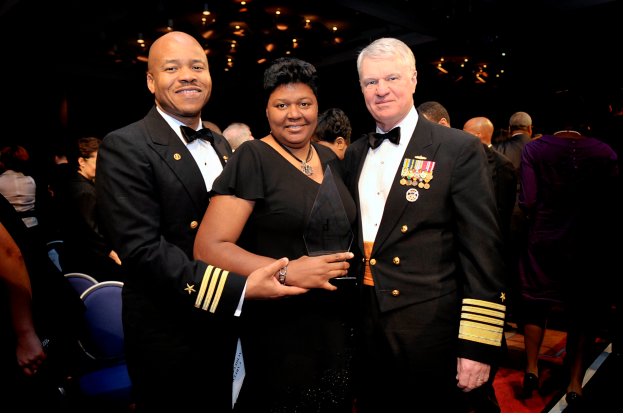 Zwei Männer und eine Frau auf einem roten Teppich, die Frau hält eine Trophäe, mit Menschen und beleuchteter Decke im Hintergrund.