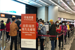 Eine Gruppe von Menschen in Masken steht in einer Schlange am Flughafen, getrennt durch Barrierepfosten mit Bändern, mit einem Schild im Vordergrund, das "Hong Kong Airport Refunds Due to Covid-19" lautet, und Säulen und Deckenleuchten im Hintergrund.