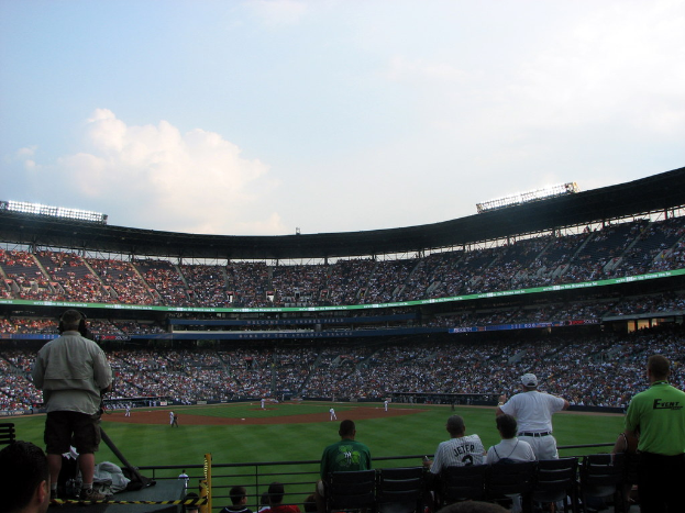 Eine Aufnahme eines Stadions mit Spielern auf einem Rasenfeld im Vordergrund und einer Gruppe von Zuschauern im Hintergrund, wobei einige Personen im vorderen unteren Bereich stehen und das Spiel beobachten.