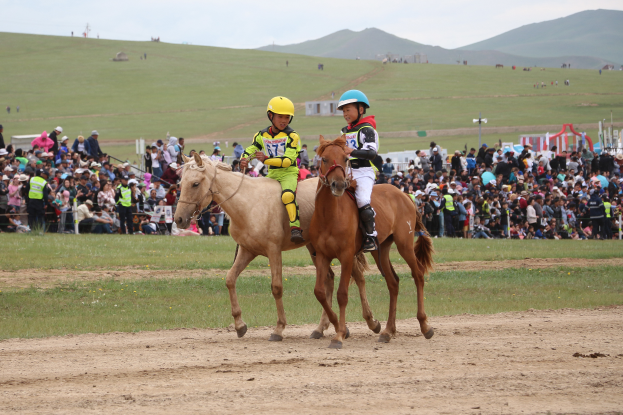 Zwei Personen mit Helmen reiten auf einem Schotterfeld, während sie von einer großen Menschenmenge im Hintergrund beobachtet werden, grasbewachsenes Gelände, entfernte Hügel und ein klarer blauer Himmel.