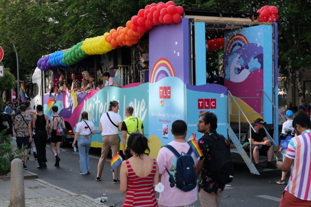 Eine Gruppe von Menschen, die auf einer Straße neben einem Lastwagen mit bunten Luftballons gehen, mit Schildern an Pfählen auf der Straße und Bäumen und Gebäuden im Hintergrund, was auf eine Pride-Parade in Paris hindeutet.