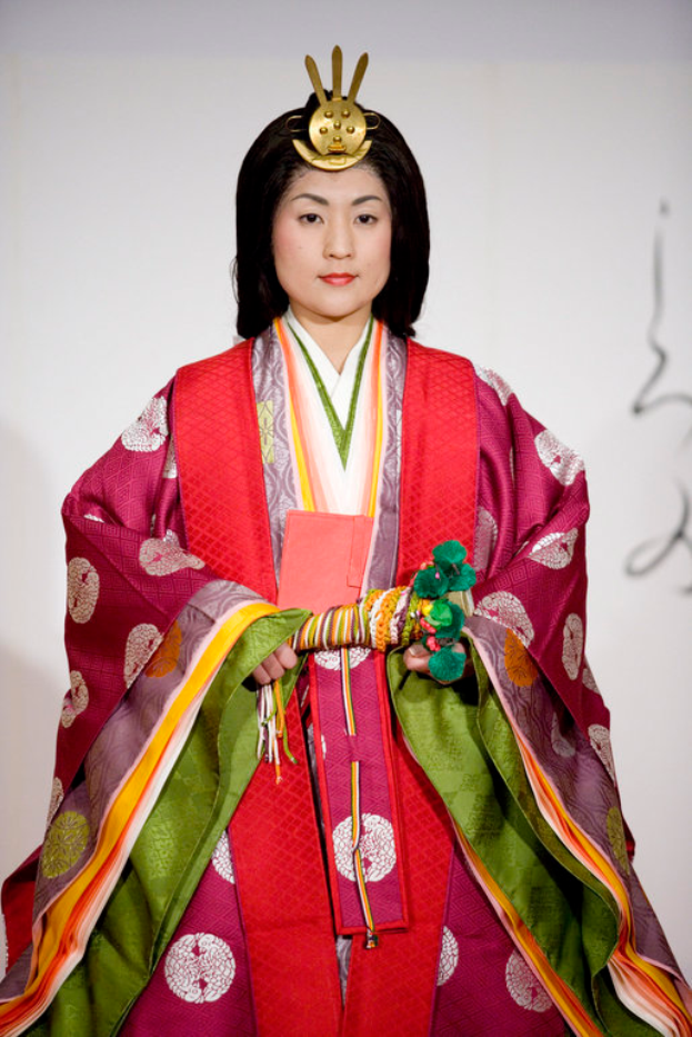 Eine Frau in einem traditionellen japanischen Kimono, das ein leuchtend rotes und grünes Kleid mit kunstvollen Mustern trägt, steht selbstbewusst auf einem Laufsteg mit ihrem Haar zu einem ordentlichen Knoten gebunden.