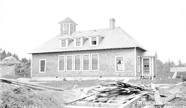 Schwarz-weiß-Foto einer Regierungsgrundschule von 1905 mit Fenstern, Türen, Bäumen und einem klaren Himmel, das hölzerne Bretter und Text am Boden zeigt.