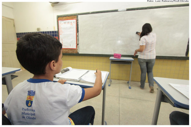 Ein Junge sitzt an einem Tisch und schreibt in ein Buch, während eine Frau die Tafel mit einem Putzlappen in einem Klassenzimmer mit mehreren Tischen, einem haltenden Kasten und einem Plakat an der Seite reinigt.
