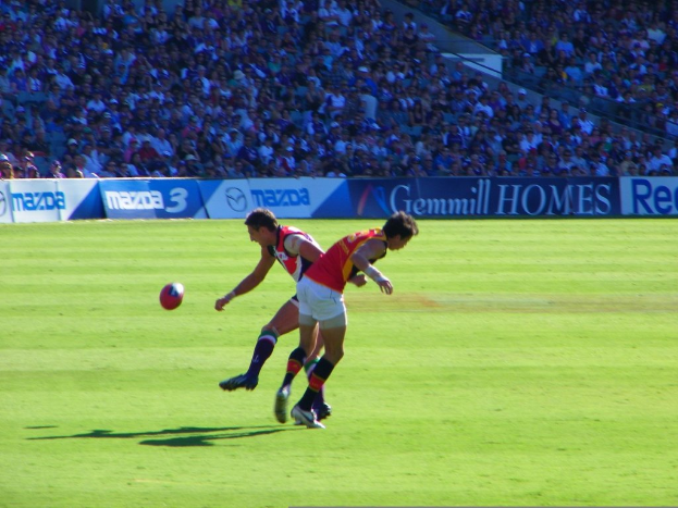 Zwei Personen in rot-weißer Kleidung stehen mit einem Ball in der Luft, während im Hintergrund eine Menge und Banner im Stadion zu sehen sind.