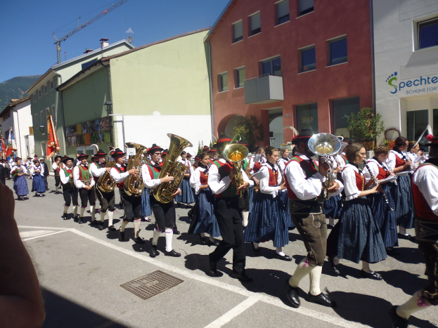 Eine Gruppe von Menschen in traditioneller bayrischer Tracht, die Musikinstrumente spielen, während sie eine Straße mit Gebäuden, Pflanzen und einem Namensschild entlanggehen, mit einem Hügel und einem klaren blauen Himmel im Hintergrund.