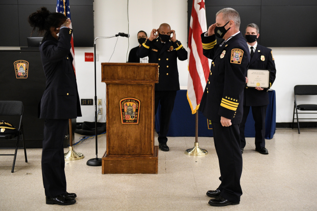 Eine Gruppe uniformierter Personen mit Masken vor einem Rednerpult bei einer Vereidigung eines neuen Feuerwehrchefs, der eine Urkunde hält, mit Fahnen, Stühlen, Tischen und einer Wand im Hintergrund.
