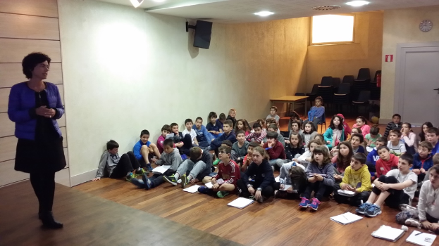 Eine Gruppe von Kindern, Jungen und Mädchen, sitzt auf dem Boden mit Büchern vor sich, während eine Frau links auf einer Bühne steht.