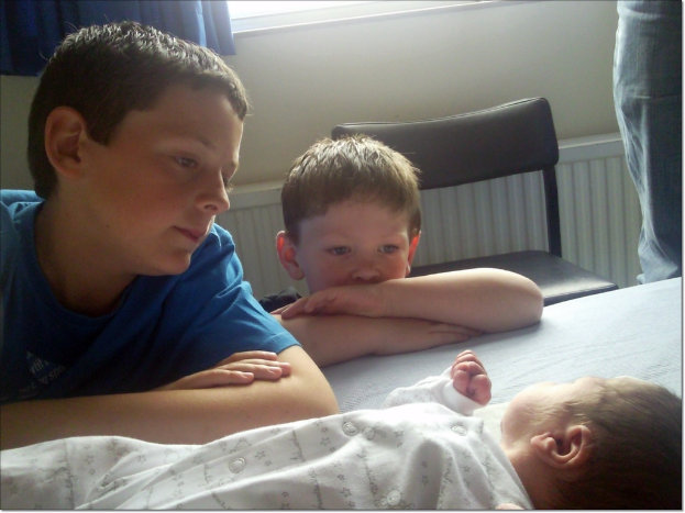 Ein Baby liegt auf dem Boden mit zwei Jungen links daneben, die das Baby anschauen, und einem Stuhl neben ihnen; ein Personenschuh ist oben rechts zu sehen, mit einer Wand und einem blauen Vorhang im Hintergrund.