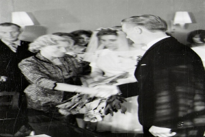 Ein Schwarz-Weiß-Foto von einer Braut und einem Bräutigam, die sich die Eheringe reichen, wobei die Braut ein weißes Kleid und eine Schleier trägt und einen Blumenstrauß hält, und der Bräutigam einen Anzug und eine Krawatte trägt, mit Wandlampen im Hintergrund.