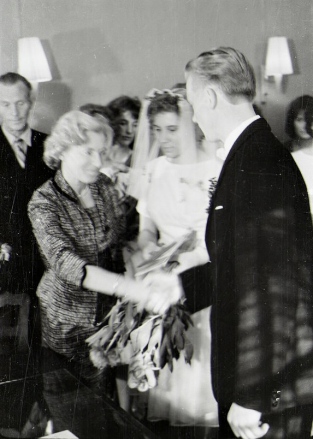 Ein Schwarz-Weiß-Foto von einer Braut und einem Bräutigam, die sich die Eheringe reichen, wobei die Braut ein weißes Kleid und eine Schleier trägt und einen Blumenstrauß hält, und der Bräutigam einen Anzug und eine Krawatte trägt, mit Wandlampen im Hintergrund.