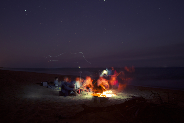 Eine Gruppe von Menschen sitzt um ein Lagerfeuer am Strand in der Nacht, mit Sand darunter und Wasser im Hintergrund, beleuchtet vom Feuer und den Sternen.