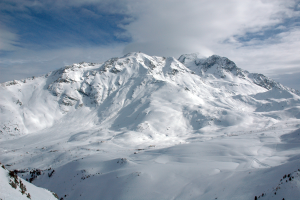 Ein schneebedeckter Berg mit ein paar Skifahrern, die darauf hinabfahren, umgeben von einem bewölkten Himmel und unberührtem Schnee.