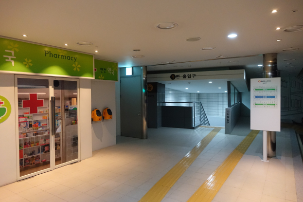 Apotheken-Eingang mit einem "Apotheke"-Schild auf der linken Seite, einer Tafel mit Text auf der rechten Seite, beleuchteten Lampen auf dem Dach und Hintergrundobjekten.