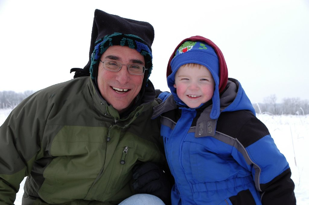 Ein Kind in einer blauen Jacke steht rechts lachend, während eine Person links sitzt und lacht, mit einer schneebedeckten Landschaft, Bäumen und Himmel im Hintergrund.