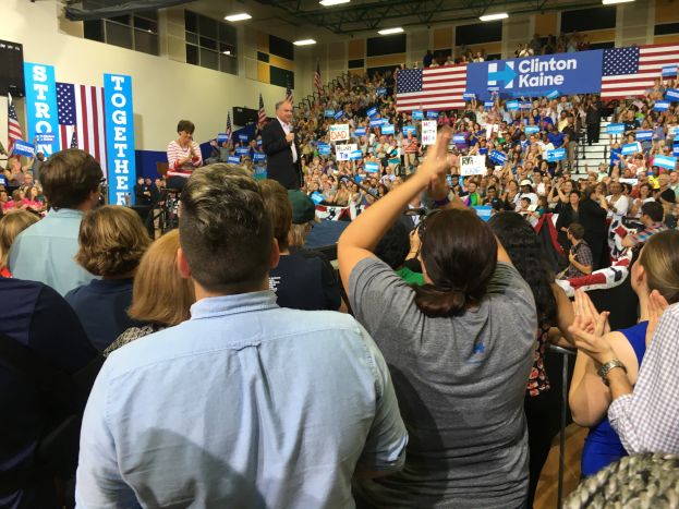 Eine große Menge steht vor einer Bühne, auf der ein Mann in ein Mikrofon spricht, mit einer Frau neben ihm, während Fahnen und Banner im Hintergrund gezeigt werden.