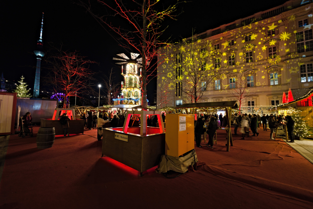 Ein lebendiger Weihnachtsmarkt in Berlin, Deutschland, mit Menschen um beleuchtete Stände, Bäume, Gebäude, Laternen und einen Turm unter einem dunklen Himmel.