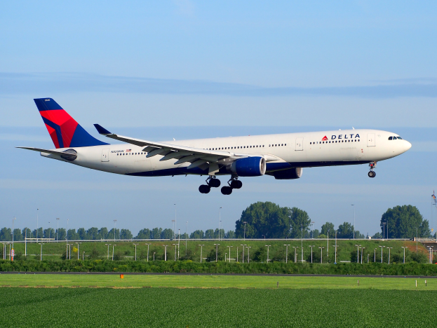 Delta Air Lines Boeing 787-9 Dreamliner beim Abheben vom Frankfurt Airport, mit grünem Gras darunter, Laternenpfählen und Bäumen im Hintergrund, einem Turm auf der rechten Seite und weißen Wolken am Himmel.