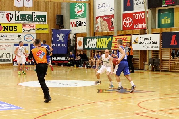 Ein Basketballfeld mit Spielern im Vordergrund und einer hölzernen Wand mit Plakaten und Bannern im Hintergrund.