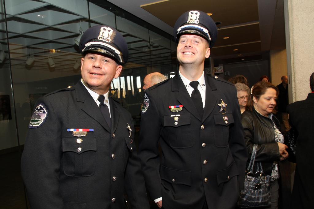 Zwei grinsende Beamte im Vordergrund, Menschen in der Mitte, Glasfenster links, Lampen und Decke oben und Säulen und Menschen rechts.